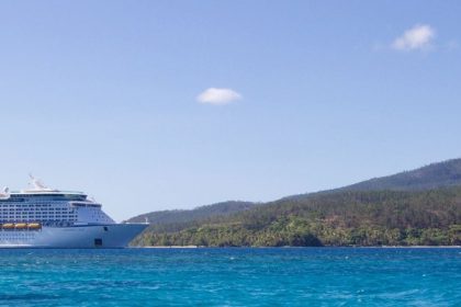 Grande navio de cruzeiro navegando próximo a uma ilha tropical, sob céu azul.