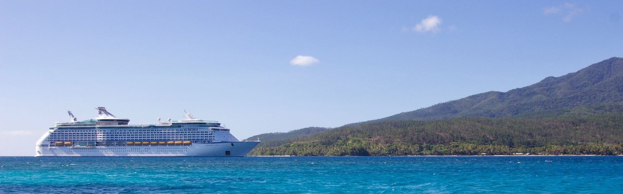 Grande navio de cruzeiro navegando próximo a uma ilha tropical, sob céu azul.